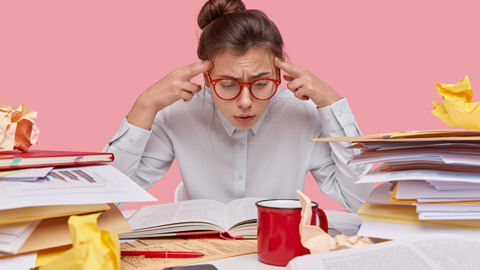 Frustrated nerd focused in book, keeps fingers on temples, tries to remember information, busy before examination session, has hair combed in bun, wears glasses, surrounded with pile of papers