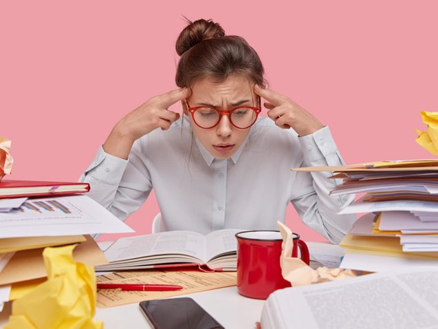 Frustrated nerd focused in book, keeps fingers on temples, tries to remember information, busy before examination session, has hair combed in bun, wears glasses, surrounded with pile of papers