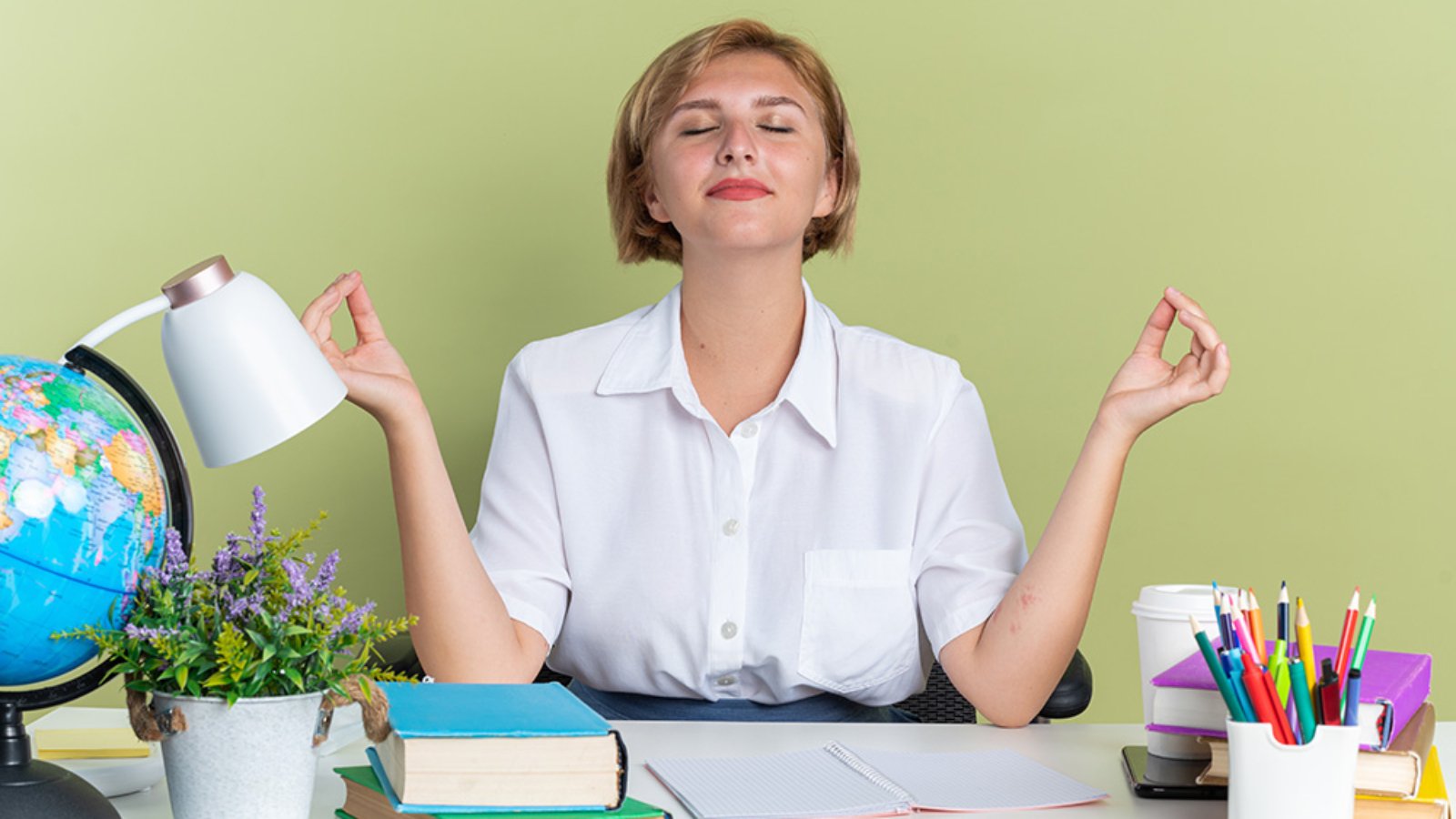 peaceful young blonde student girl sitting at desk with school tools meditating with closed eyes isolated on olive green background