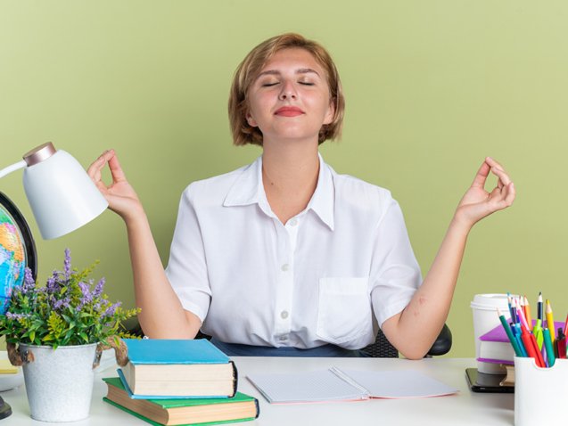 peaceful young blonde student girl sitting at desk with school tools meditating with closed eyes isolated on olive green background